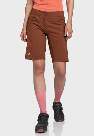 Braune Shorts mit einem schlanken Design, ausgestattet mit zwei Knöpfen, Seitentaschen und einem dezenten Logo. Kombiniert mit rosa Socken und schwarzen Fahrradsportschuhen.