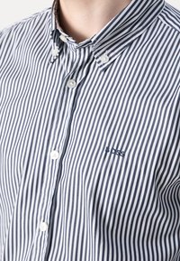 Gros plan d'une personne portant une chemise à rayures verticales noir et blanc, boutonnée avec un col boutonné et "BOSS" brodé sur la poitrine.