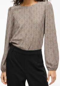 Ilgas rankovėmis šviesiai ruda palaidinė su tekstūruotu gėlių raštu ir pūstomis rankovėmis, derinama su juodomis pritaikytomis kelnėmis.