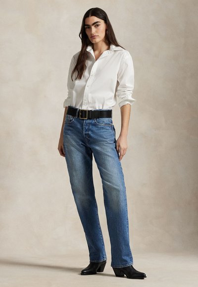 Noor naine kannab valget nööbitavat särki, siniseid teksapükse musta vöö ja musta kannaga saapaga, seisab neutraalse tausta ees.