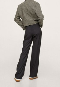 Personne portant une veste à carreaux marron et un pantalon large sombre, se tenant les mains sur les hanches, devant un fond clair uni.