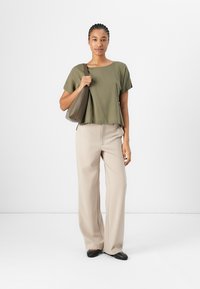 Top corto verde oliva abbinato a pantaloni beige a gamba larga. Tiene in mano una borsa marrone testurizzata e indossa scarpe nere flat, il tutto su uno sfondo bianco.