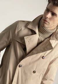 Gabardina beige de doble botonadura con grandes botones negros, un cuello ancho y una tela suave y ligera, combinada con un suéter beige acanalado.
