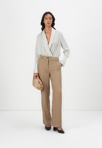 Hvit bluse med prikker, beige tilpassede høyhalsbukser, holder en beige clutch, kombinert med svarte spisse flate sko.
