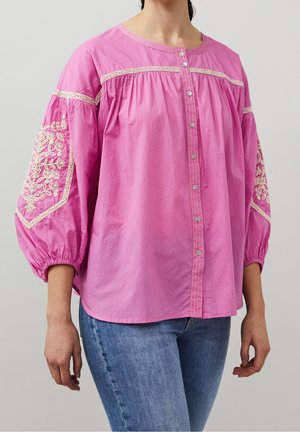 Femme portant un chemisier rose ample avec des motifs floraux brodés sur les manches bouffantes et un jean bleu sur un fond uni.