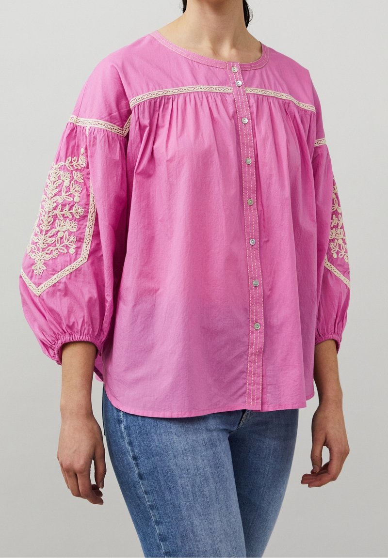 Femme portant un chemisier rose ample avec des motifs floraux brodés sur les manches bouffantes et un jean bleu sur un fond uni.