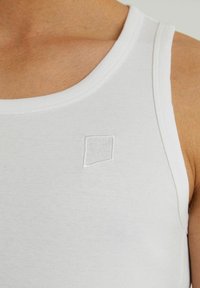 Bela bombažna majica brez rokavov s prilegajočim krojem, ki ima majhen bel vezen kvadratni logotip na levi strani prsnega predela. Gladka tekstura.