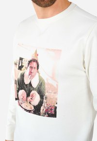 Vit sweatshirt med en färgglad grafik av en karaktär i grön outfit med en fluffig krage, som håller en tallrik med mat.