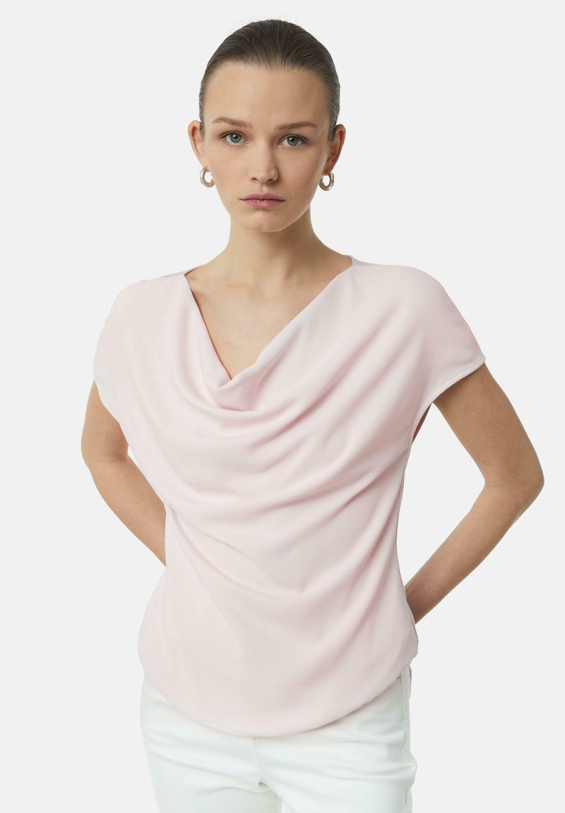 Helles rosa Drapiertes Blusenshirt mit weitem Ausschnitt und kurzen Ärmeln, aus einem glatten Stoff gefertigt. Das Model trägt weiße Hosen und hat die Hände in die Hüften gestützt.