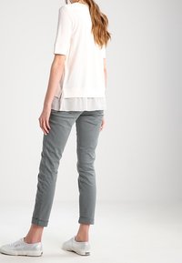 Jasna kremowa bluzka z krótkim rękawem, nałożona na przeźroczysty panel, zestawiona z dopasowanymi szarymi spodniami i srebrnymi trampkami. Prosty, czysty design w całym zestawieniu.