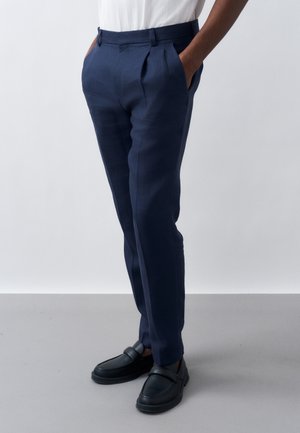 Άτομο που φοράει μπλε-ναυτικό παντελόνι ραμμένο στα μέτρα με τα χέρια στις τσέπες και μαύρα μοκασίνια χωρίς κορδόνια, στέκεται μπροστά σε απλό φόντο.