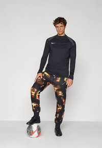 Musta pitkähihaurheilupaita harmailla yksityiskohdilla, yhdistetty värikkäisiin, abstraktikuosisiiin housuihin ja mustiin jalkapallokenkiin, seisomassa pallon päällä.