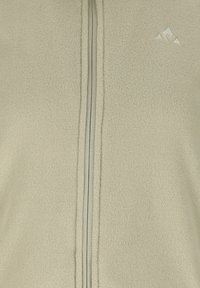 Lys beige fleece-stof med central lynlåslukning og en lille trekantet broderet logo øverst til højre.