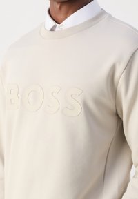Ljust beige sweatshirt med en texturerad "BOSS"-logotyp. Klassisk crew neck-design med slät tyg och subtil glans.