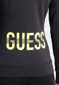 Haut de sport noir avec un grand texte jaune "GUESS" au dos, au design ajusté et à la texture de tissu lisse.