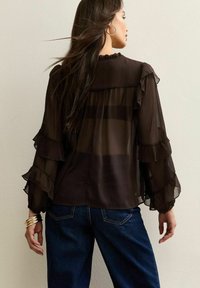 Zwarte doorzichtige blouse met gerimpelde mouwen en een gegolfde halslijn, gelaagd ontwerp, gecombineerd met donkere denim jeans en metalen armbanden.
