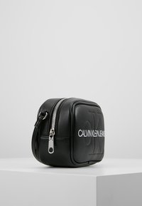 Czarna skórzana torba na ramię ma prostokątny kształt, zamknięcie na zamek, wytłoczone logo oraz elementy w kolorze srebrnym.