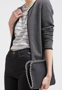 Texturiert schwarzer Cardigan mit Rundhalsausschnitt, getragen über einem grau-weißen gestreiften Hemd. Eine schwarze Clutch mit Kettendetails liegt in der Hand.