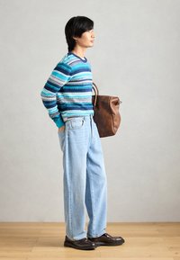 Pull rayé dans des tons bleus et verts, jean rétro clair, sac en cuir marron et chaussures foncées, se tenant devant un fond neutre.