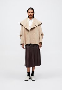 Beige, oversized gyapjú keverék kabát széles gallérral, dupla gombolású kialakítással, barna bőr midi szoknya és fehér slip-on cipők.