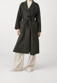 Abrigo largo de lana verde oliva con una cinturilla, solapas de pico y bolsillos laterales, llevado sobre pantalones de pierna ancha de canalé color crema y zapatos blancos.