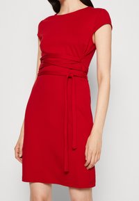 Czerwona bawełniana sukienka z krótkimi rękawami i okrągłym dekoltem. Posiada owinięty pas z możliwością regulacji, gładką fakturę oraz długość do kolan.