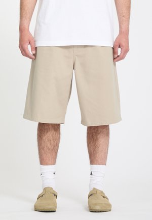 Homme portant un short beige jusqu'aux genoux, des chaussettes blanches, des chaussures beige à enfiler et un t-shirt blanc, debout devant un fond uni.