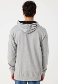 Sudadera gris con capucha ajustable, mangas largas, puños y dobladillo de canalé, con un corte relajado y una textura suave. Vista desde atrás.
