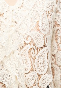 Tissu en dentelle blanche avec des motifs floraux et cachemire complexes, présentant des bords festonnés et un fond en filet semi-transparent.