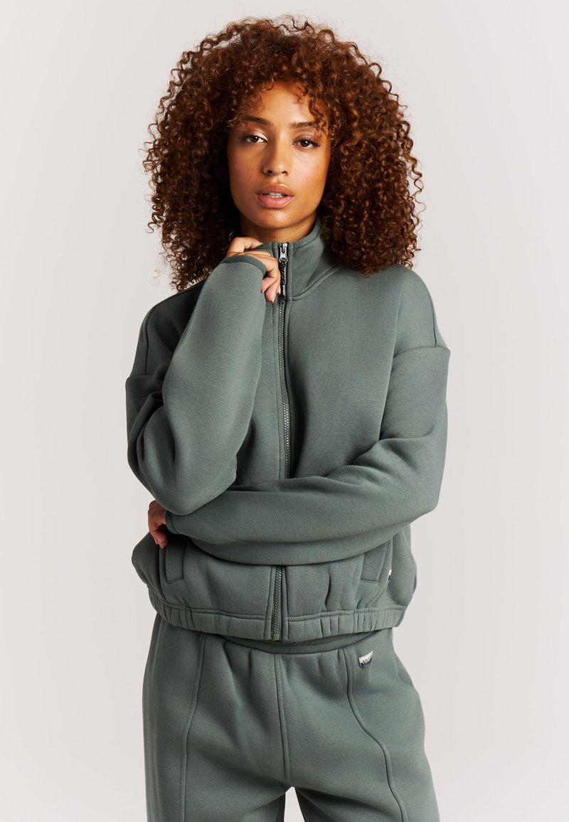 Vrouw met krullend haar die een grijs-groene hoodie met rits en bijpassende broek draagt, poserend met één hand bij haar kin.