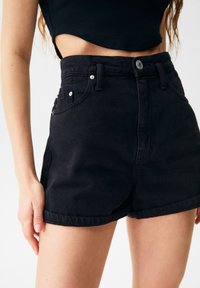 Černé džínové šortky s vysokým pasem mají našitý lem, přední kapsy a zapínání na kovový knoflík, s jemnou texturou a přiléhavým střihem.