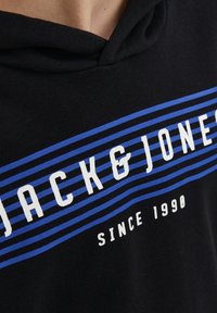 Sudadera negra con rayas horizontales azules y texto blanco que dice "JACK & JONES" con "SINCE 1990" debajo. Tela suave, diseño informal.