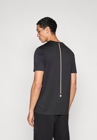 Melnā sporta krekls ar īsām piedurknēm, kas atrodas vertikāla zelta rāvējslēdzēja detaļa muguras daļā un teksturēts auduma akcents.