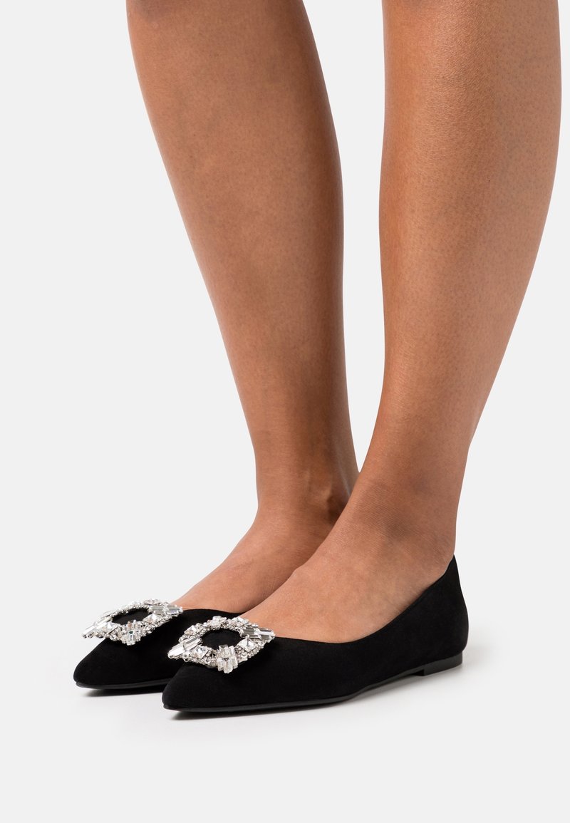 Ballerines en daim noir avec un bout pointu et une bouclerie ornée de strass sur le cou-de-pied. Semelle plate.