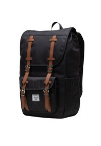Zwarte stoffen rugzak met bruine leren riemen, gespen, voorvak en een wit rechthoekig logo-label op het onderste voorvak.