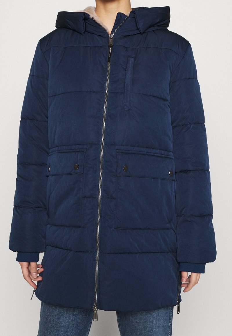 Manteau matelassé bleu marine avec une fermeture éclair intégrale, de grandes poches avant et une capuche. Le matériau présente une texture lisse avec des coutures horizontales.