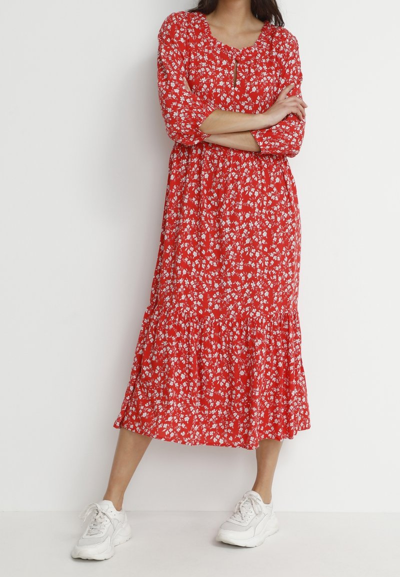 Femme portant une robe midi rouge à fleurs avec des manches trois-quarts et des baskets blanches, debout les bras croisés et une jambe légèrement en avant.