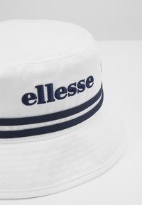 Cappello a secchiello bianco in tela, con nome del marchio ricamato in blu e due strisce blu attorno alla base. Texture liscia e forma strutturata.