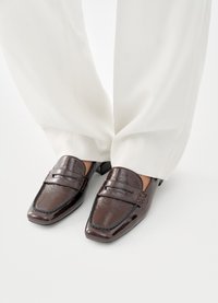 Καφέ μοκασίνια από δερματίνη με τετράγωνη μύτη, λεπτομερείς ραφές και χαμηλό τακούνι, σε συνδυασμό με λευκό παντελόνι φαρδιάς γραμμής.