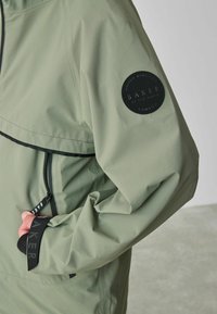 Groene waterdichte jas met een zwart ronde patch met de tekst "BAKER," een ritszak en elastische accenten op de mouwmanchetten.