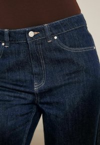 Donkerblauwe denim jeans met een klassieke pasvorm, zilveren hardware en contrastgele stiksels langs de zakken en naden.