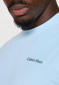 hellblaues, kurzärmliges Hemd aus glattem Stoff mit einem schwarzen "Calvin Klein"-Logo auf der Brust. Einfaches Halsdesign.