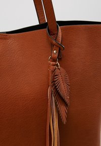 Bruine leren tote bag met een gestructureerde afwerking, voorzien van decoratieve franjes met bladaffiches en een zwarte voering. Inclusief verstelbare schouderband.