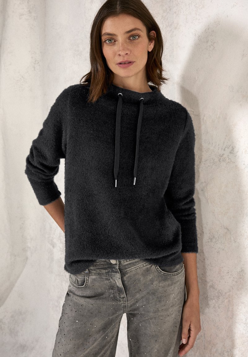 Svart, fluffig hoodie med dragsko; har långa ärmar och en avslappnad passform. Matchas med grå jeans prydda med nitar.