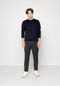 Pull-over en maille à manches longues de couleur marine associé à un pantalon sombre et des baskets blanches. Le pull présente des poignets et un col côtelés, avec une texture lisse.