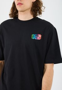 Black cotton t-shirt featuring a colorful graphic resembling a ticket on the left chest, with text and a star design in vibrant shades.