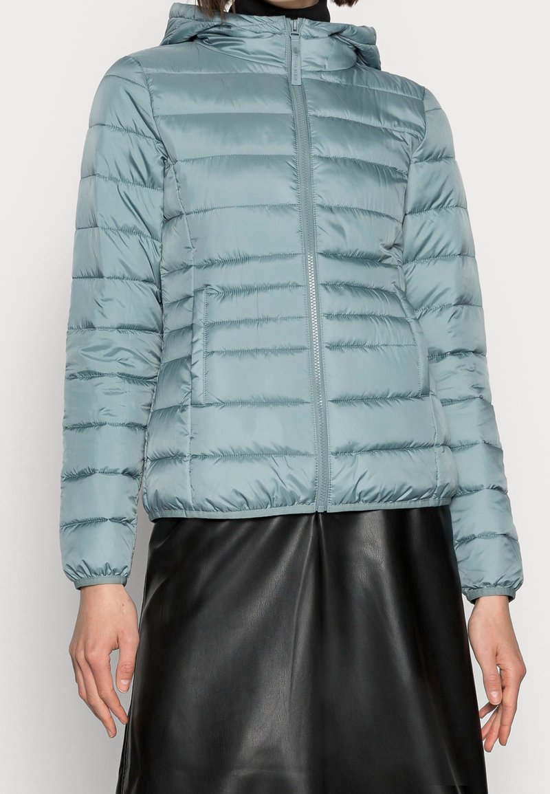 Personne portant une veste matelassée à capuche bleu clair zippée, associée à une jupe en cuir lisse noire, sur un fond uni.