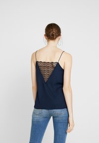 Mörkblå tanktopp med spetsdetalj i V-form på ryggen och tunna axelband, tillverkad av mjukt material. Matchas med ljusblå denimjeans.