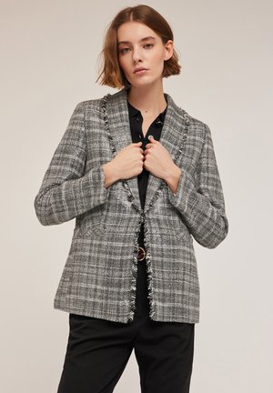 Szary blazer w kratę z postrzępionymi brzegami, zapinany na pojedynczy guzik, z klapami w stylu notch. Noszony na czarnej koszuli, z przednimi kieszeniami i dopasowanym krojem.
