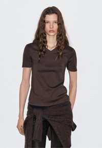 T-shirt marrone a V con maniche corte, realizzato in tessuto liscio, indossato con un avvolgente scuro e texture. Design semplice con una forma aderente.